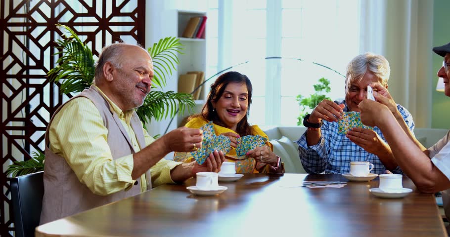 Indian senior adults playing cards game while seated at dining table in cozy modern home, laughing and bonding joyfully, Asian old aged friends celebrating cheerful indoor reunion with fun activity