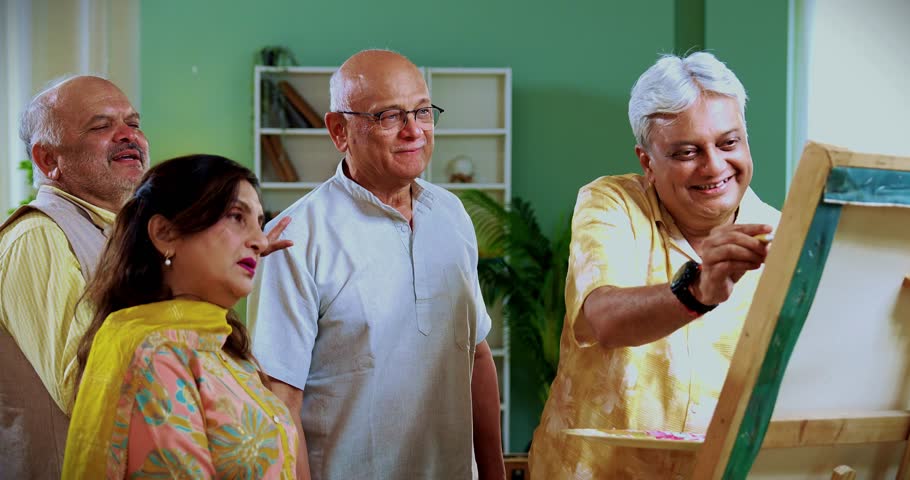 Senior man paints while friends praise or laugh at his art, Indian Asian old aged group enjoying creative fun indoors as retired male paints with brush and canvas during cozy home reunion gathering
