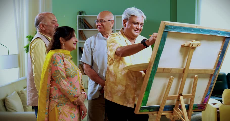 Senior man paints while friends praise or laugh at his art, Indian Asian old aged group enjoying creative fun indoors as retired male paints with brush and canvas during cozy home reunion gathering