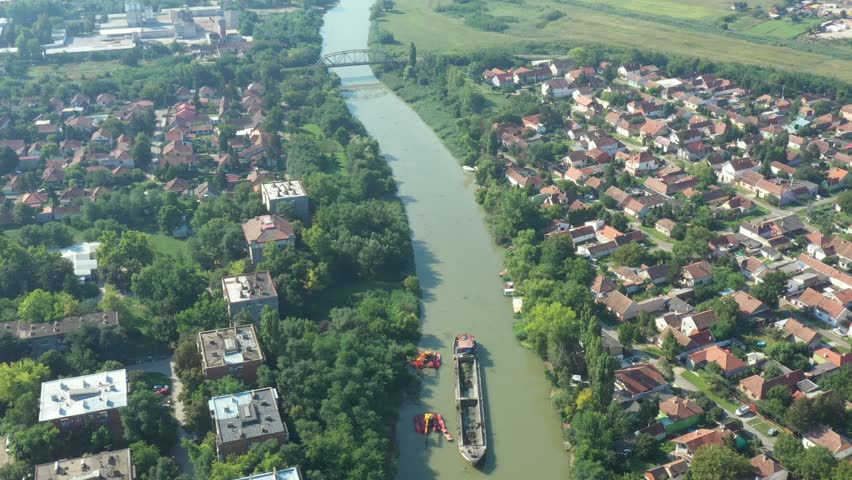 Above view over two excavators dredge as they dredging, working on river, canal, deepening and removing sediment, mud from riverbed in a polluted waterway. Dolly move panning on settlement, city