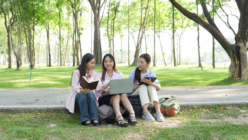 Group of college students with laptops and tablet computers doing homework in a park, talking and working together under a tree outdoors.