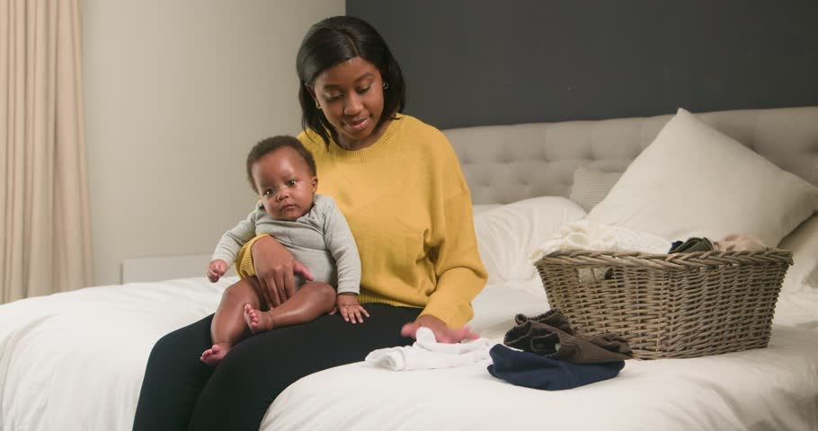 Young Mother Folds Laundry While Holding Her Baby on Bed: A Heartwarming Scene of Family Life and Motherhood