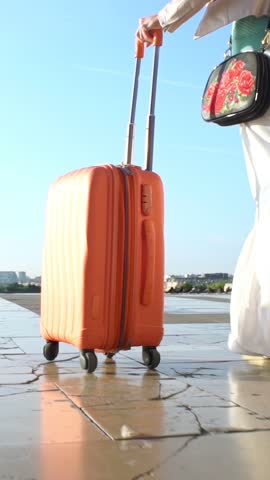 Woman rolling orange suitcase in sunlight. Excitement of travel, the freedom of departure, and the anticipation of a new destination. Escape, change, and the beginning of a new story.