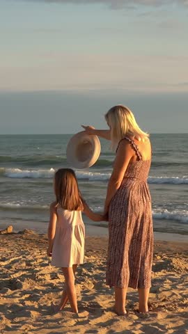 Back view of mother and daughter stand and look at sea on beach at sunset. Summer vacation travel holiday. People walk outdoors. Vertical shot