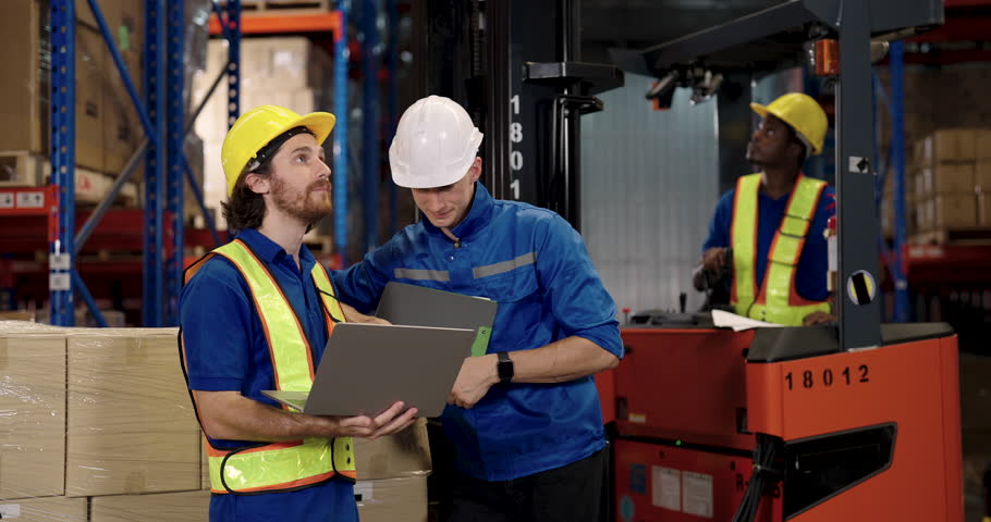 Warehouse supervisor standing next to forklift explaining work procedures to staff using laptop checking report inside organized industrial storage space during logistics team coordination effort
