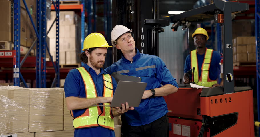 Warehouse supervisor standing next to forklift explaining work procedures to staff using laptop checking report inside organized industrial storage space during logistics team coordination effort