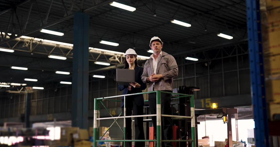 Caucasian male engineer and Asian female supervisor stand on elevated platform inside warehouse pointing upward while discussing inventory process during slowmotion equipment inspection session