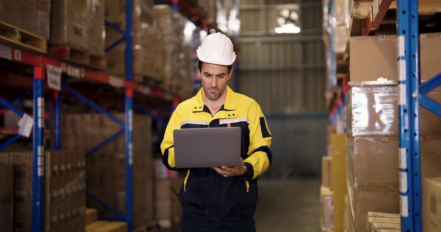 Caucasian male warehouse worker standing in aisle using laptop to check inventory stock while taking short break from packaging process inside modern industrial logistics center