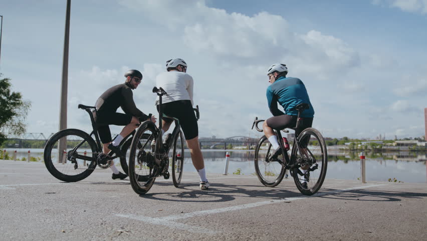 Group of bicyclists with helmets rests looking at calm river from city embankment. Sportsmen with professional vehicles prepare for race on riverbank rear view