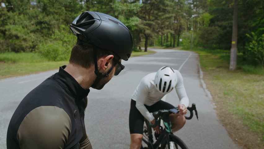 Sportsman wearing helmet shares water with friend resting on country road. Professional cyclists participate in long distance racing across green forest