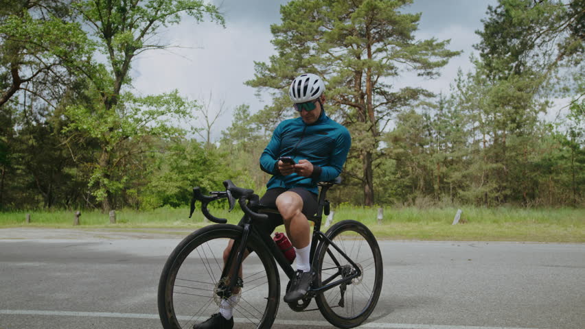 Man rider wearing helmet texts on smartphone sitting on bicycle at summer countryside. Sportsman surfs internet on cellphone resting during bike training