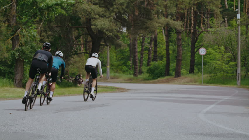 Trained athletes in sportswear ride bicycles along curved road. Athletic men group on professional bikes competes on racing at green countryside slow motion