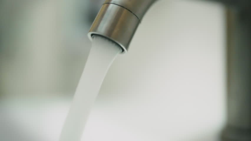 A stainless steel faucet releases a steady stream of water, creating a soft splash in a bright kitchen environment. The sunlight enhances the clean atmosphere, reflecting off the surfaces