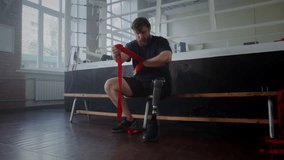 Bearded male athlete with prosthetic leg uses hand wrap sitting near boxing ring. Young boxer with disability and sports equipment prepares for fight in gym - Powered by Shutterstock - Get 15% off with code: PIKWIZARD15