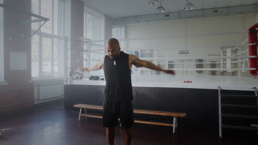 Black bearded man fighter rotates arms warming up shoulder joints near boxing ring. Focused African American athlete exercises before fighting in gym
