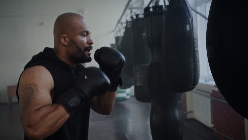 Focused African American man boxer beats heavy punching training in gym. Black bald sportsman practices strong punches with boxing equipment in fighting club