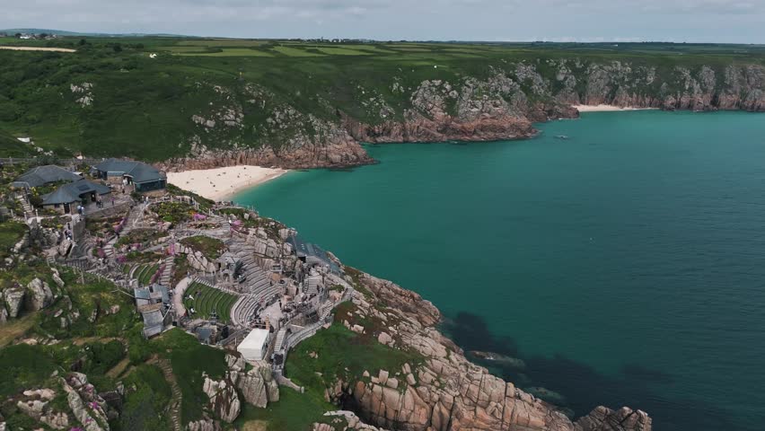 Rightward drone orbit shows Minack Theatre terraces and dramatic cliff edge by the sea, aerial establishing overview