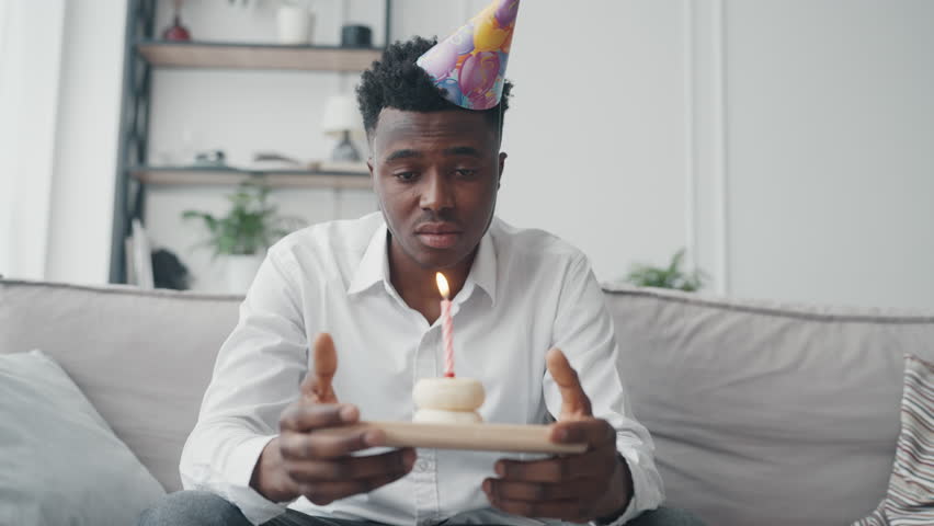 Unhappy male person spending his birthday party alone, make wish and blowing candle, vertical video. Celebrating anniversary without friends and family, stay alone and suffering, lonely black guy