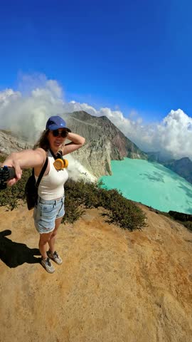 Selfie perspective of a woman standing on the rim of a volcano crater with a turquoise acidic lake and sulfur fumes behind her at Ijen volcano, Indonesia.
