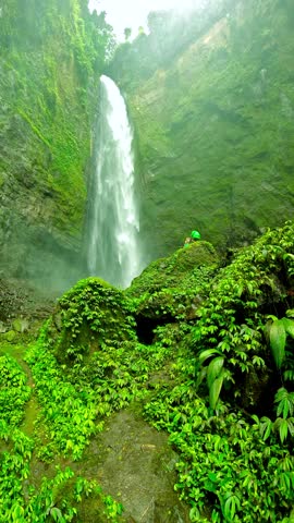 Waterfall flows down a lush green cliff face with mist and a person at the base, Kabut Pelangi Waterfall, East Java, Indonesia.