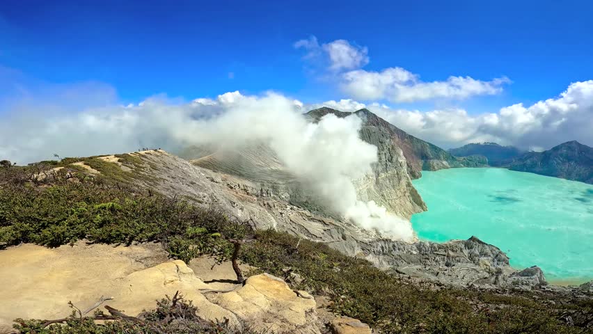 View across the rocky ridge of Ijen volcano caldera showing steam rising and the turquoise acidic crater lake, East Java, Indonesia.