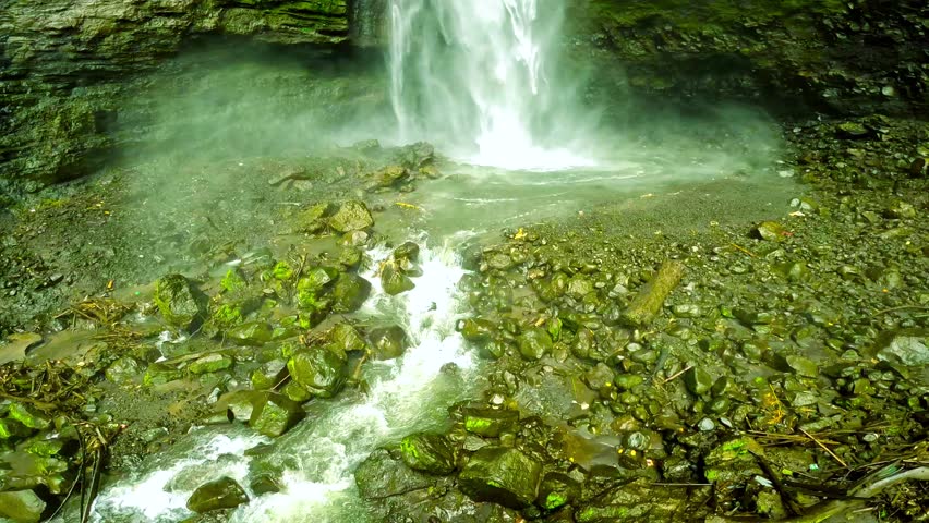 Waterfall cascading down mossy rocks into a rocky pool. Lush green cliff face surrounds the falling water. Located in East Java, Indonesia.