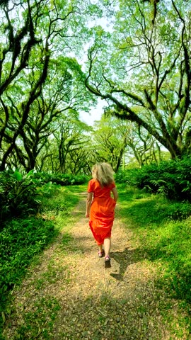 Woman in an orange dress runs down a path through a mystical forest with ancient gnarled trees, De Djawatan Forest, Indonesia.