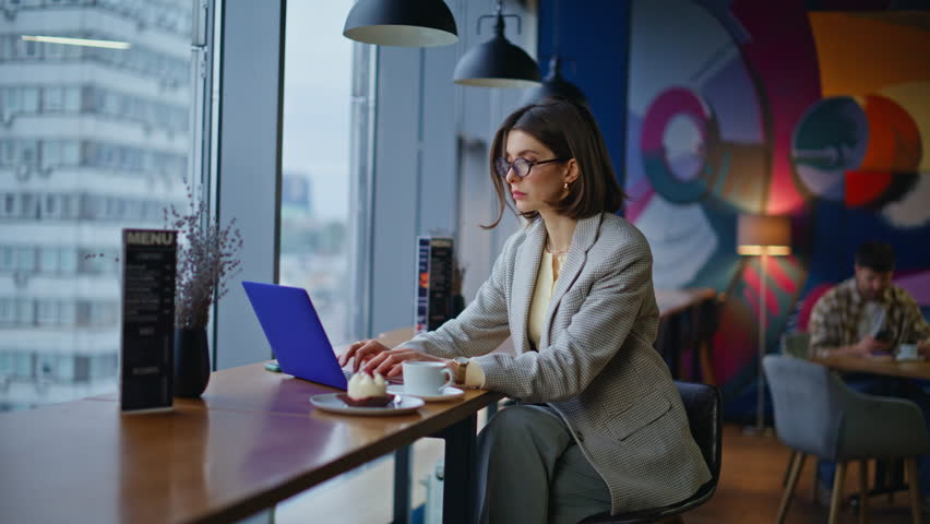 Eyeglasses smm specialist browsing laptop in coffee shop thinking on creative project closeup. Serious woman work online in bright cafe. Happy brunette manager typing remote job feeling successful