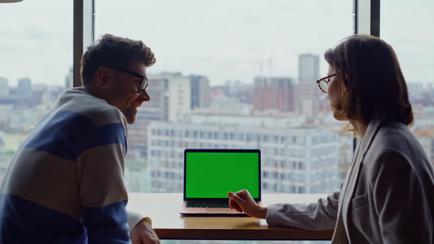 Two employees working chroma key device in glass walls office rear view. Closeup two business people chatting mockup laptop in open space. Colleagues pointing green screen computer speaking together 
