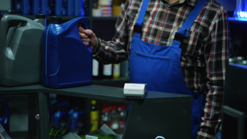 Customer using a smartphone for contactless payment at a terminal while buying motor oil from a senior auto parts store clerk.