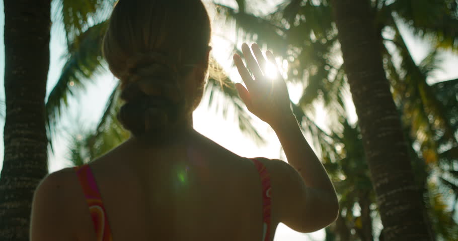 Woman raises hand to block sun while standing under palm trees on tropical beach