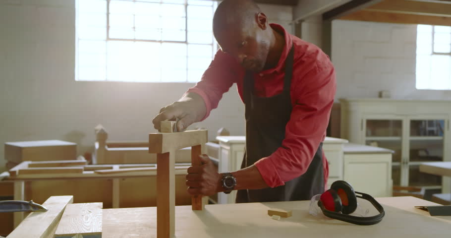 African American man placing frame then sanding beam with block at workbench for smooth finish. Craftsman, woodworking, craftsmanship, workshop, furniture, carpentry, manual labor
