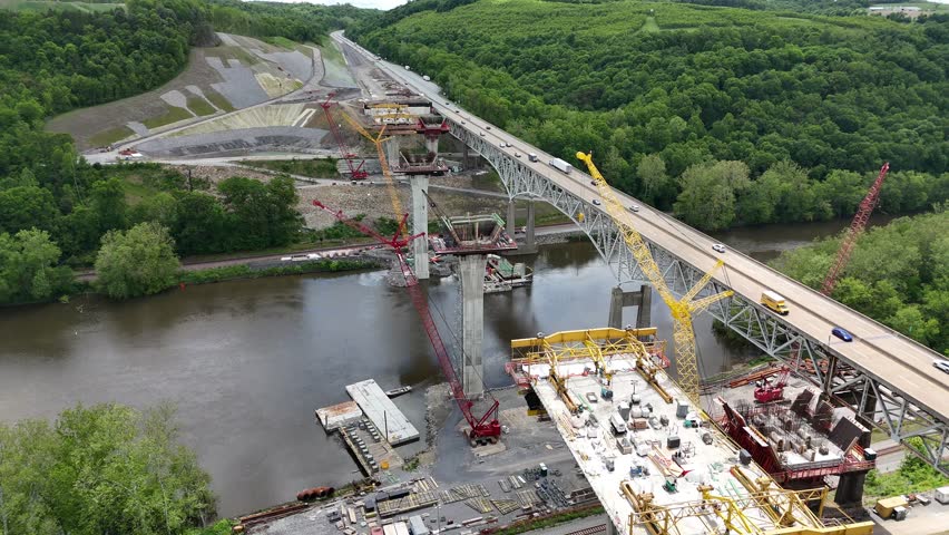 Aerial view of highway I-80 in Pennsylvania showing a bridge over a river surrounded by lush forests and mountains, with heavy traffic and massive construction of a second highway bridge nearby.