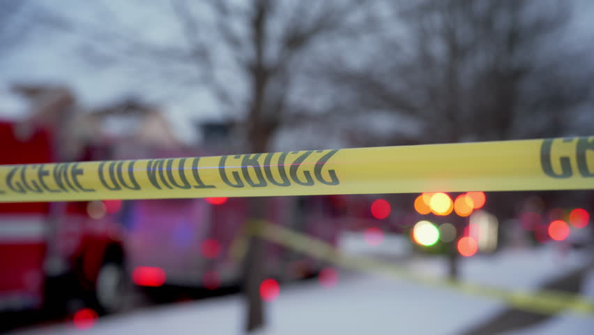 A bright yellow Crime Scene Do Not Cross tape stretched across the street at residential area, marking a restricted area for investigation. Flashing lights are on the background