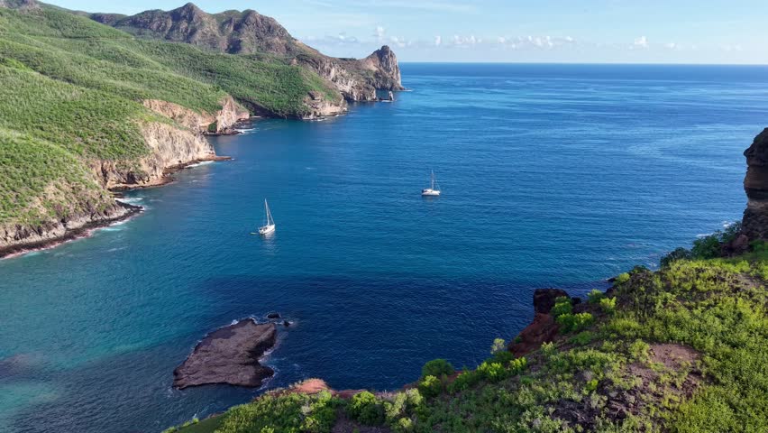 Drone pans across a beautiful landscape of a remote anchorage with sailboats. Towering cliffs soar above the crashing ocean below. 
