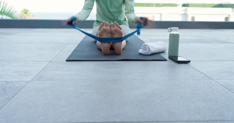 Grasping blue band Asian woman pulling inward for hamstring stretching on balcony mat with towel. Fitness, flexibility, wellness, relaxation, outdoor, minimalism, rejuvenation