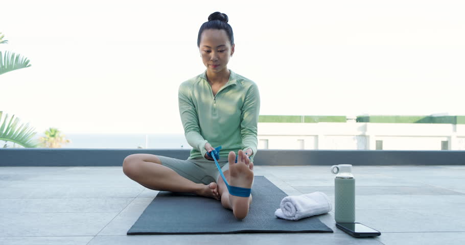 Grasping blue band Asian woman pulling inward for hamstring stretching on balcony mat with towel. Fitness, flexibility, wellness, relaxation, outdoor, minimalism, rejuvenation