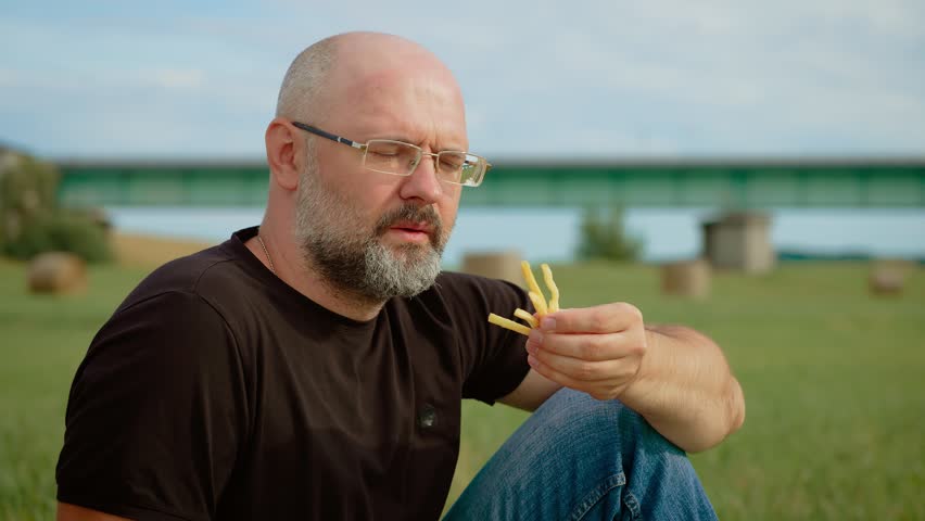 Confident man eating fries in agricultural field, unhealthy food. Determined male snacking on French fries while relaxing in rural field. Assured guy enjoying fast food amidst peaceful countryside