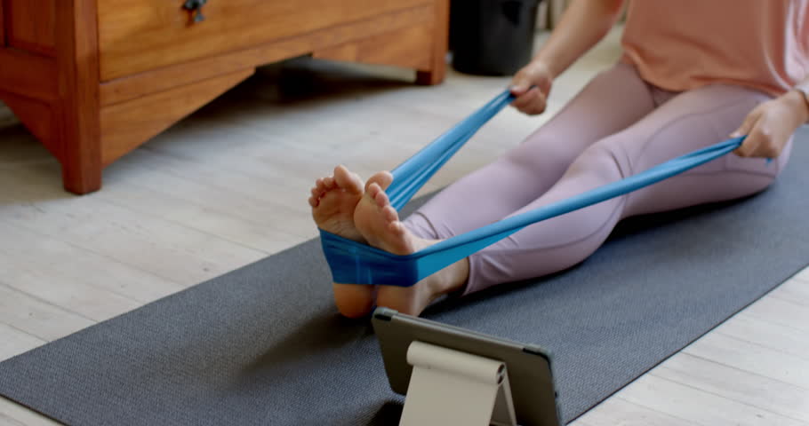 Asian woman following tablet workout pulling band and checking smartwatch to track progress at home. Fitness, strength, flexibility, mindfulness, wellness, home gym, sport