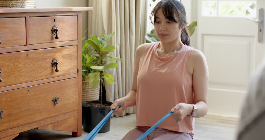 Asian woman following tablet workout pulling band and checking smartwatch to track progress at home. Fitness, strength, flexibility, mindfulness, wellness, home gym, sport