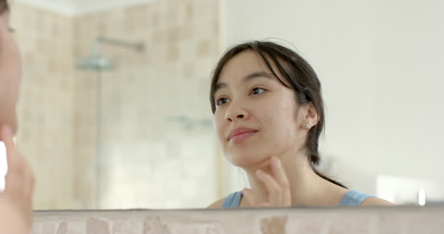 Asian woman massaging moisturizer on cheeks at bathroom mirror, adjusting bangs, wearing smartwatch. Skincare, grooming, selfcare, beauty, reflective, wellness, minimalism