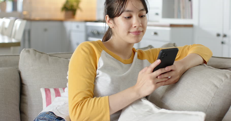 Receiving phone notification, Asian woman reclining on grey sofa scrolling, smiling and responding. Casual, cozy, modern, lifestyle, relaxation, communication, home