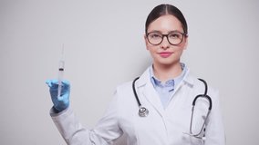 Smiling female doctor in white lab coat and gloves poised with medical syringe, representing vaccination or medical treatment, against light white background - Powered by Shutterstock - Get 15% off with code: PIKWIZARD15