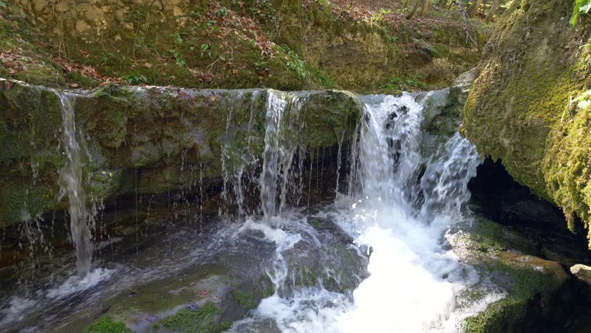 A scenic footage of a small waterfall surrounded with algae-covered rocks in the woods on a sunny day