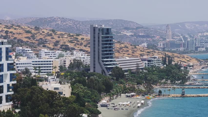 Aerial view of Limassol, Cyprus, with the Mediterranean Sea, breakwaters, boats, modern buildings, and rolling hills in clear sunlight.