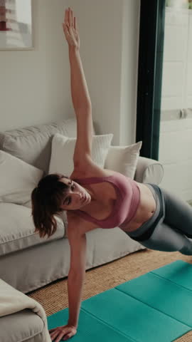 Vertical video, Young woman doing yoga and pilates exercises in her living room at home on a mat. She performs a side plank pose, looking healthy and peaceful.