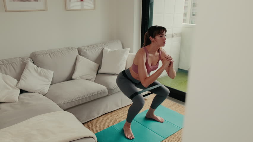 A young woman performs squats with resistance band in her living room. She follows online fitness videos shown on TV, enjoying a home workout session.