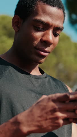 Vertical video, A focused Black man checks his fitness app on his phone outdoors. He analyzes his workout progress and prepares for his next exercise.