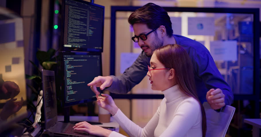 Two Asian software engineers collaborate at night in modern office, writing machine learning algorithms and debugging artificial intelligence code on multiple monitors in AI tech development workflow.