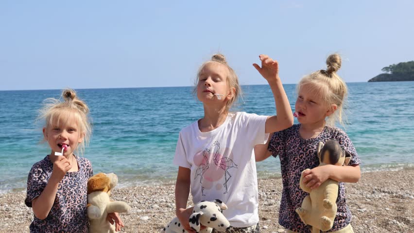Three little triplet sisters are standing on the sea beach in summer with toys in their hands